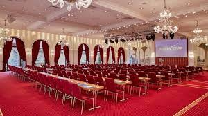 Salle de conférence avec sièges rouges, tables, écran, lustres et décorations murales.