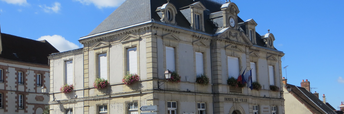 Mairie à deux étages avec toits en ardoise, fenêtre à balustrades, fleurs aux fenêtres, voiture blanche devant.