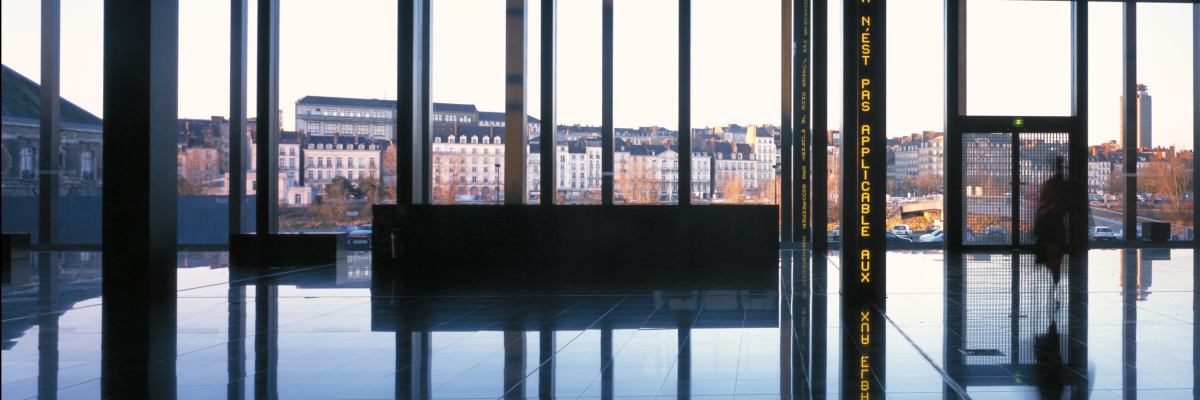 Salle avec grandes fenêtres, sol réfléchissant, banc central et vue sur des bâtiments.