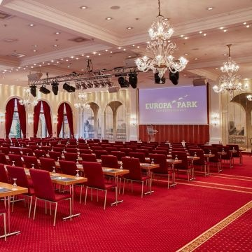 Salle de conférence avec chaises, tables, écran, projecteur, lustres et rideaux.