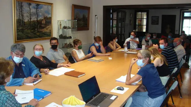 Réunion avec participants masqués autour d'une table, ordinateurs portables, documents, tableaux et peinture murale.