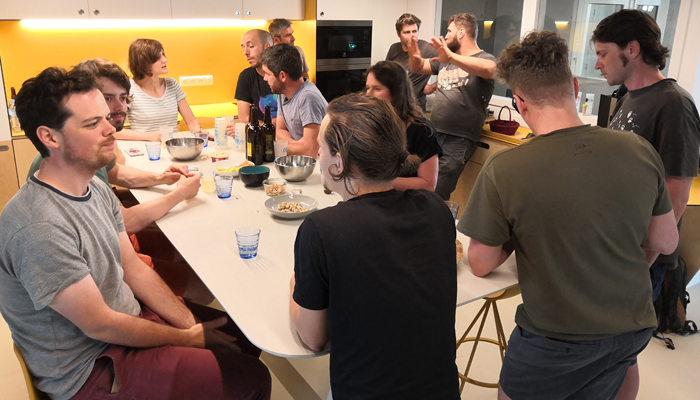 Groupe de personnes assis et debout autour d'une table dans une cuisine, avec des bouteilles, des verres, des assiettes et de la nourriture sur la table.