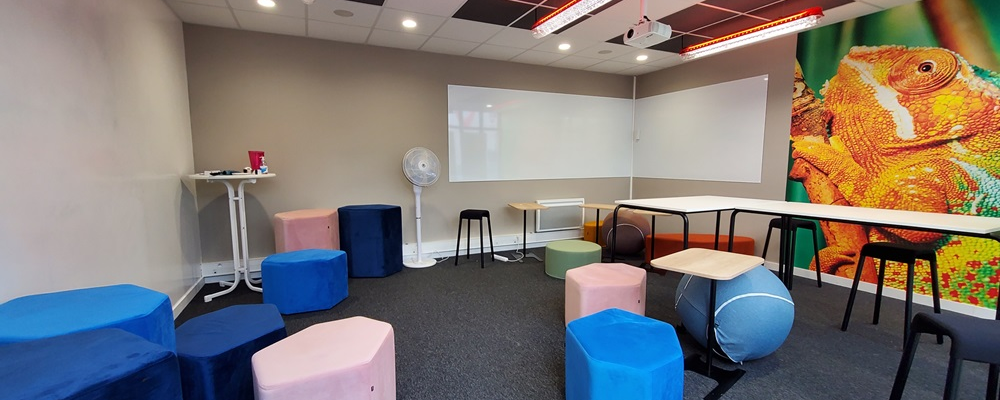 Salle avec écran, vidéoprojecteur, chaises, tables, coussins et mur mural.