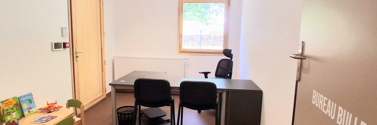 Bureau avec table, chaises, radiateur, fenêtre, porte, petit bureau avec livres et jouets, panier de recyclage, speakers.