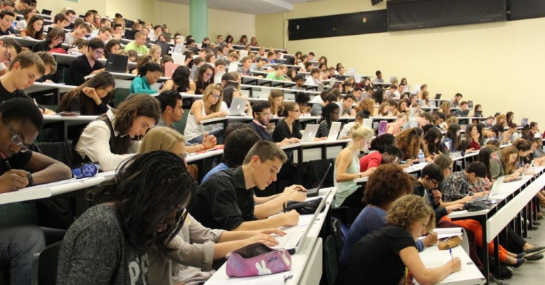 Amphithéâtre rempli d'étudiants avec ordinateurs portables, tables et écrans.