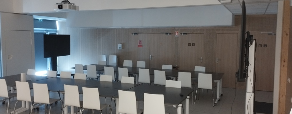 Salle de classe avec tables, chaises, écran, vidéoprojecteur et tableau blanc.
