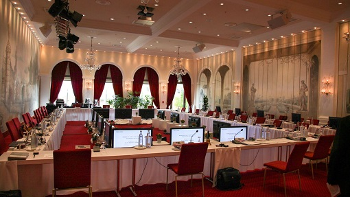 Salle avec tables, chaises, écrans, projecteurs, plantes et rideaux.