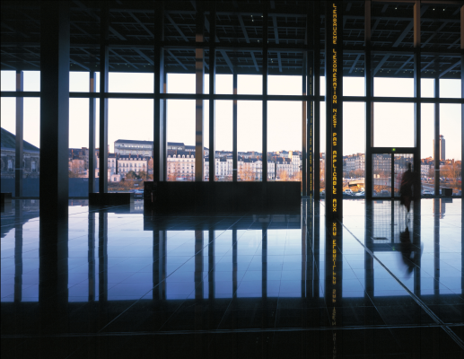 Salle avec grandes fenêtres, sol réfléchissant, banc central et vue sur des bâtiments.