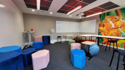 Salle avec écran, vidéoprojecteur, chaises, tables, coussins et mur mural.